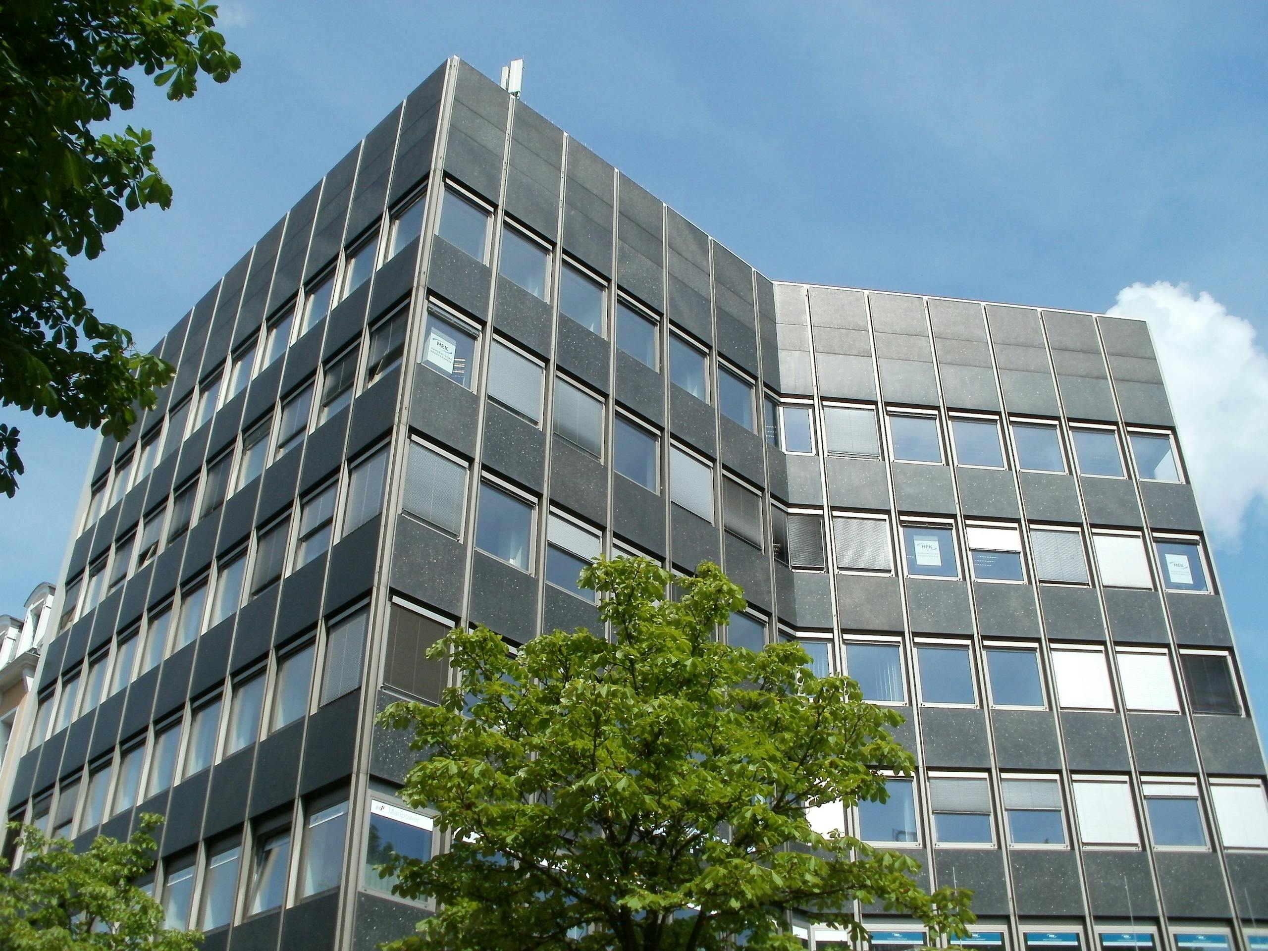 Low angle view of a modern office building against a clear blue sky, framed by green trees.